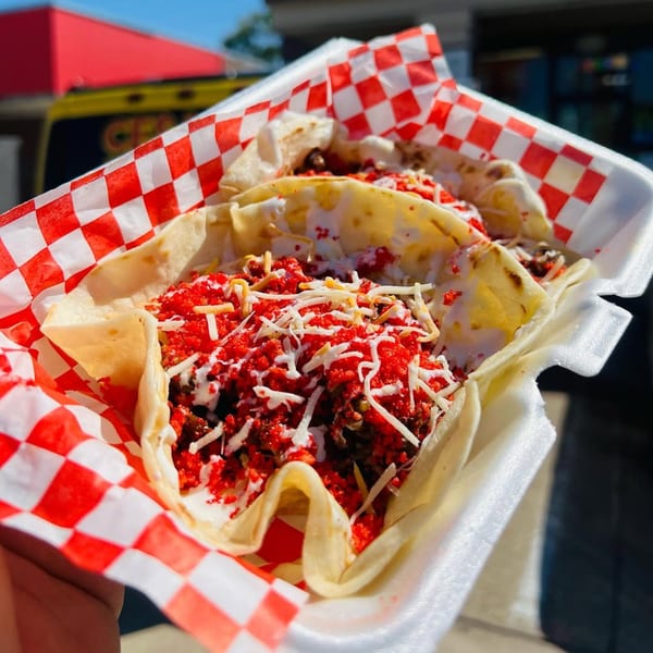 Two Tex-Mex tacos with red crunchy toppings, white sauce, and shredded cheese in a red-checkered basket.