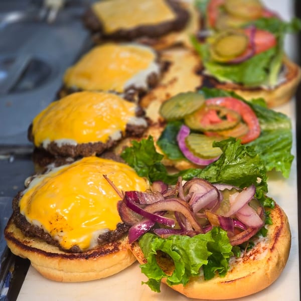 Row of cheeseburgers with melted cheese, lettuce, pickles, tomatoes, and grilled onions on buns.