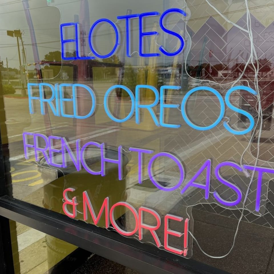 Neon signs in a window advertise "Elotes," "Fried Oreos," "French Toast," and "& More."