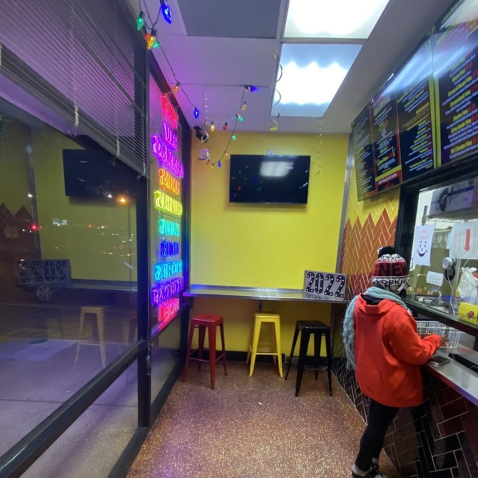 A person in a red hoodie orders at Cesar's Snack Shack, with colorful neon signs and string lights.