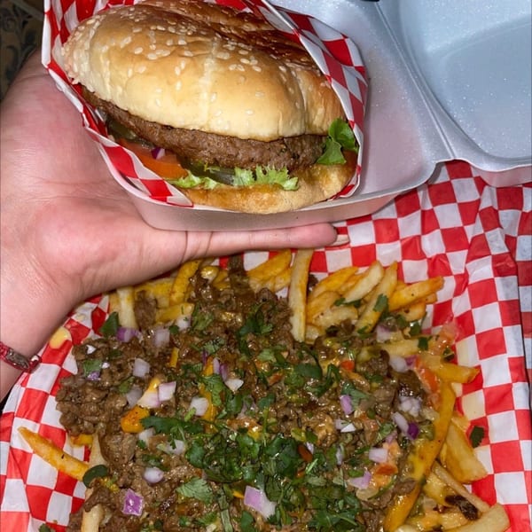A hand holds a burger in a red and white checkered wrapper above a basket of carne asada fries.